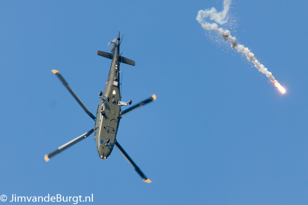 Ein militärisches Helikopter in der Luft, das eine Rakete hält, die Rauch und Feuer ausstößt, mit Text in der linken unteren Ecke.