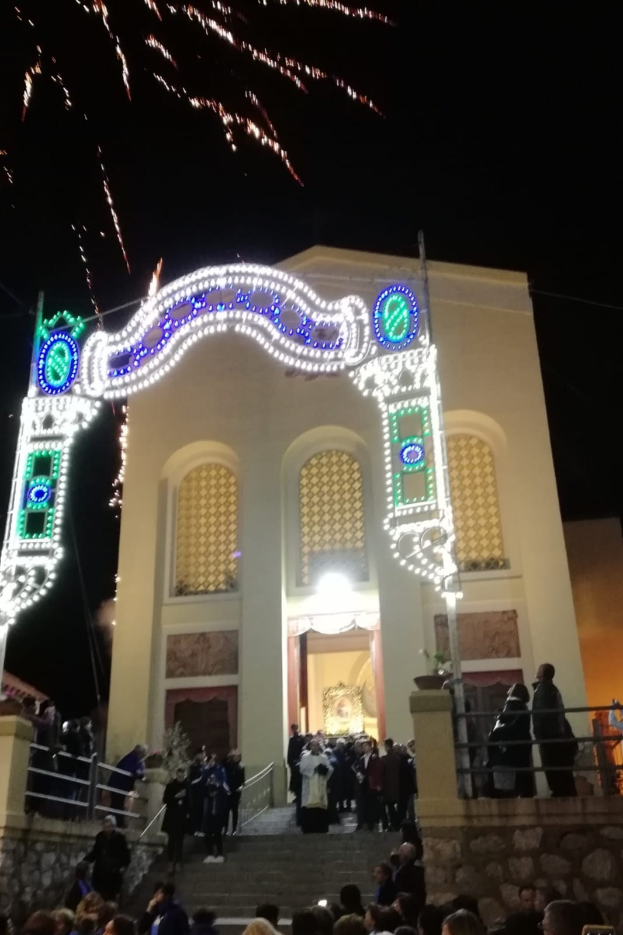 Eine Gruppe von Menschen steht vor einem Gebäude mit einem Bogen, der mit Lichtern geschmückt ist, Treppen mit Geländern und Töpfen mit Pflanzen, mit einer Feuerwerksdarbietung, die den Himmel im Hintergrund erhellt, während einer Silvesterfeier.