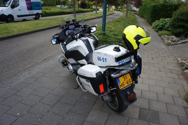 Eine Polizeimotorrad auf der Straße geparkt mit einem Helm darauf, umgeben von Bäumen, Pfählen, Gebäuden, Pflanzen, Gras, Steinen und anderen Fahrzeugen im Hintergrund.