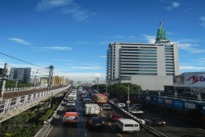 Stadtstraße mit starkem Verkehr, hohen Gebäuden, einer Brücke auf der linken Seite, Strommasten mit Drähten, Bäumen und einem bewölkten Himmel.