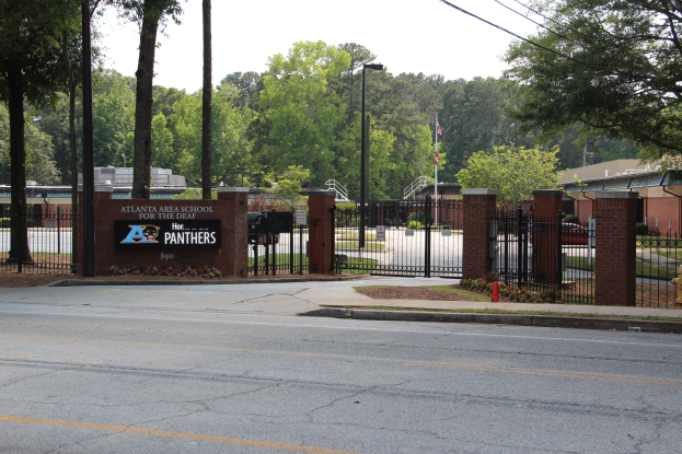 Schuleingang mit einem Schild, umgeben von Bäumen, Straßenlaternen, Fahrzeugen und Gebäuden unter einem klaren blauen Himmel.
