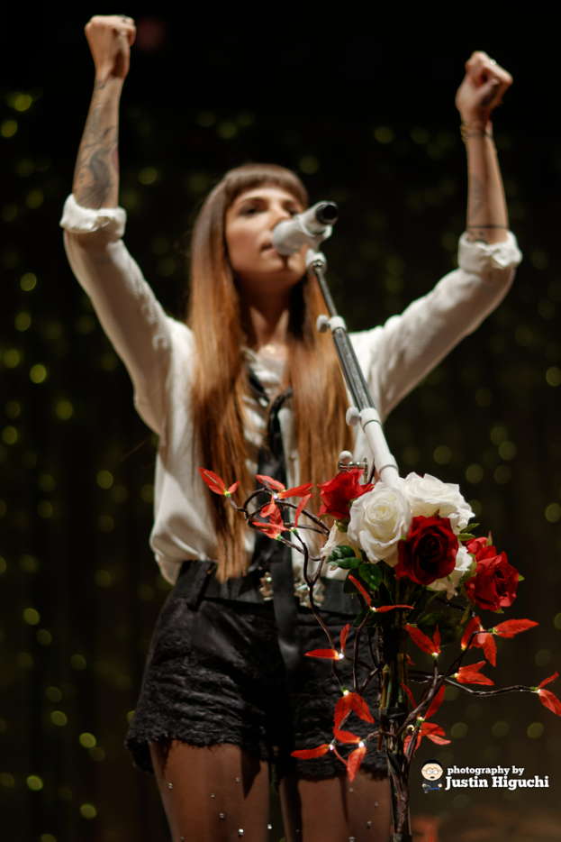 Eine Frau in einem weißen Hemd und schwarzen Shorts steht an einem Mikrofon mit Blumen im Vordergrund und spricht.