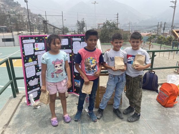 Gruppe von Kindern auf einem Basketballfeld mit Papiertüten in der Hand, mit Vorräten auf dem Boden und einer Tafel mit Text links daneben, vor einer Außenkulisse mit Zaun, Pfosten und Gebäuden.