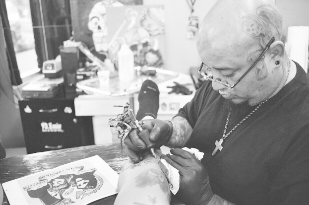 Ein Mann in schwarzem T-Shirt, Brille und einer Kette um den Hals tätowiert ein Bein mit einer Maschine, mit einem Papier auf der Oberfläche und einem unscharfen Hintergrund mit einer Flasche, Tasse und anderen Gegenständen.