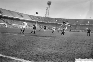 Eine Gruppe von Männern, die Fußball auf einem Feld mit Stadion, Flutlicht und klarem Himmel im Hintergrund spielen.