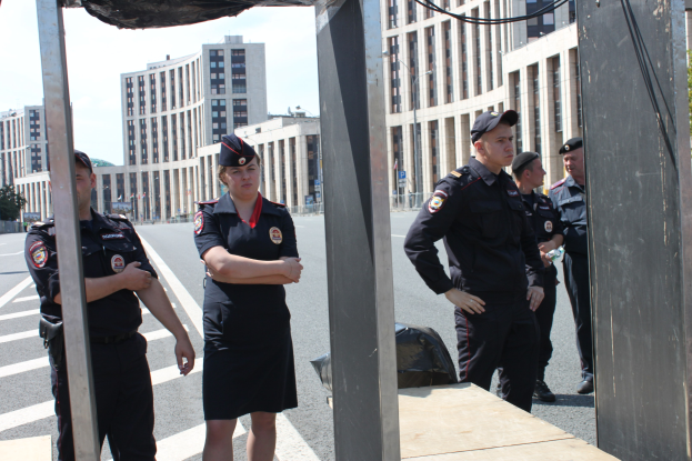 Eine Gruppe von Polizisten in Uniform, die nebeneinander auf einer Straße stehen, mit Masten und Drähten im Vordergrund, Gebäuden, Bäumen und einem klaren blauen Himmel im Hintergrund.