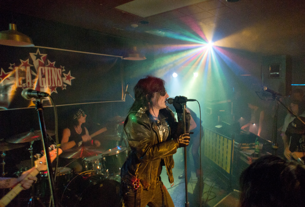 Eine Frau in einer schwarzen Jacke singt in ein Mikrofon auf der Bühne, mit einem Mann in Sonnenbrille und einem Stirnband, der ein Instrument spielt, und einem Plakat mit der Aufschrift "LA Guns" im Hintergrund, unter Diskoleuchten und hängenden Lichtern.