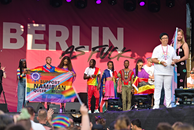 Gruppe von Menschen auf einer Bühne mit einem Banner, auf dem 'Berlin as Fuck' steht, mit Lautsprechern, Mikrofonstativen und einer feiernden Menge unten.