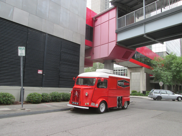 Ein Van steht in der Mitte des Bildes mit einem Auto auf der rechten Seite, und Gebäude sind im Hintergrund sichtbar.