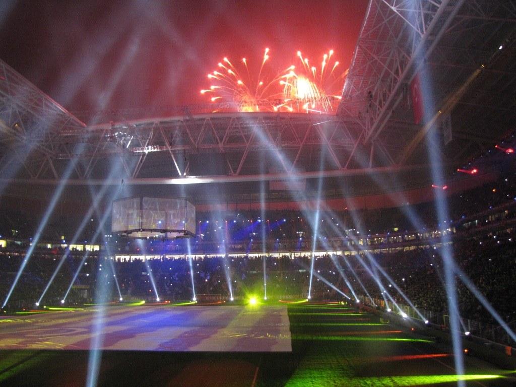 Olympisches Stadion in London bei Nacht, erleuchtet von hellen Lichtern und Feuerwerken, mit einem großen Bildschirm im Vordergrund und einer Menge drinnen.