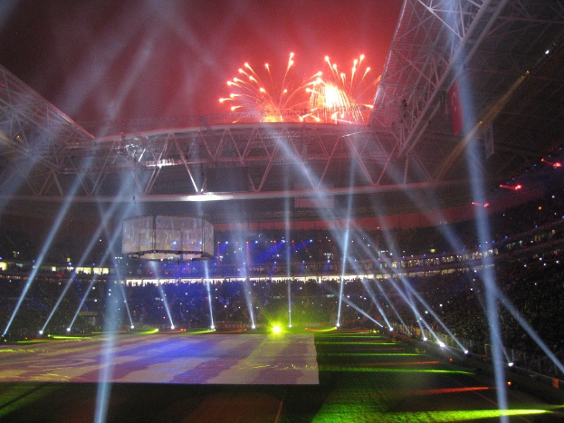 Olympisches Stadion in London bei Nacht, erleuchtet von hellen Lichtern und Feuerwerken, mit einem großen Bildschirm im Vordergrund und einer Menge drinnen.