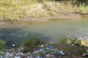 Ein Haufen Müll an einem Flussufer, umgeben von saftigem Grün und Pflanzen, mit kristallklarem Wasser im Hintergrund.