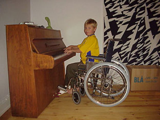 Ein Junge in einem gelben Hemd und Hose sitzt in einem Rollstuhl und spielt auf einem Piano mit Spielzeugen darauf, neben einem Vorhang und einer Kiste auf dem Boden.