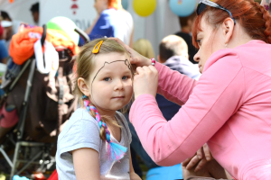 Ein junges M"dchen in einem grauen T-Shirt mit einem Pferdeschwanz l"sst sich von einer Frau auf einem Festival das Gesicht schminken, mit Menschen, Ballons, einem wei{\ss}em Zelt und einem Kinderwagen mit Gegenst"anden im Hintergrund.
