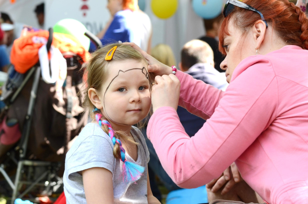 Ein junges M"dchen in einem grauen T-Shirt mit einem Pferdeschwanz l"sst sich von einer Frau auf einem Festival das Gesicht schminken, mit Menschen, Ballons, einem wei{\ss}em Zelt und einem Kinderwagen mit Gegenst"anden im Hintergrund.