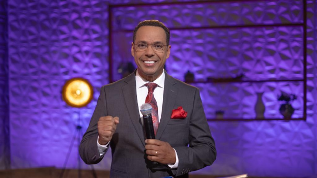 Stephen Colbert, Moderator von *The Daily Show mit Trevor Noah*, steht auf einer Bühne und hält ein Mikrofon in der Hand, trägt einen Anzug, Krawatte und eine Brille, während im Hintergrund ein Scheinwerfer und ein Ständer zu sehen sind.