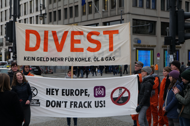 Gruppe von Menschen mit einem Banner "Divest Hey Europe, Don't Frack Us" auf einer Straße mit Gebäuden, Straßeninfrastruktur, Fahrzeugen, Bäumen und einem klaren Himmel im Hintergrund.