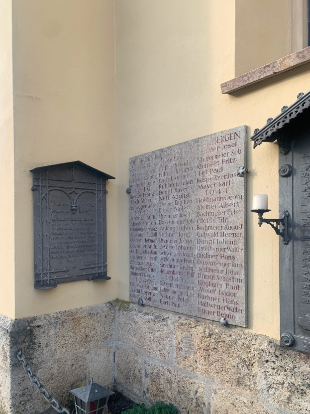 Ein Gebäude mit einer Holocaust-Gedenktafel, einem Fenster, einer Tür, einer Leuchte, einer Kette und umgebenden Pflanzen.