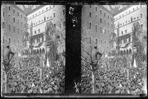 Ein Schwarz-Weiß-Foto einer großen Menschenmenge vor einem Gebäude mit Fenstern, Bäumen und Flaggen im Hintergrund, die scheinbar eine Protestveranstaltung abhalten, bei der einige Schilder und Transparente gehalten werden.