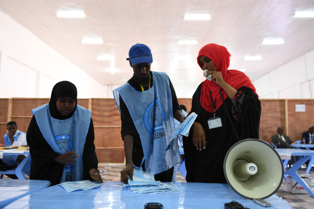Menschen versammeln sich um einen Tisch mit Megafon und Papieren an einer Wahllokal, mit anderen im Hintergrund unter beleuchteter Decke.