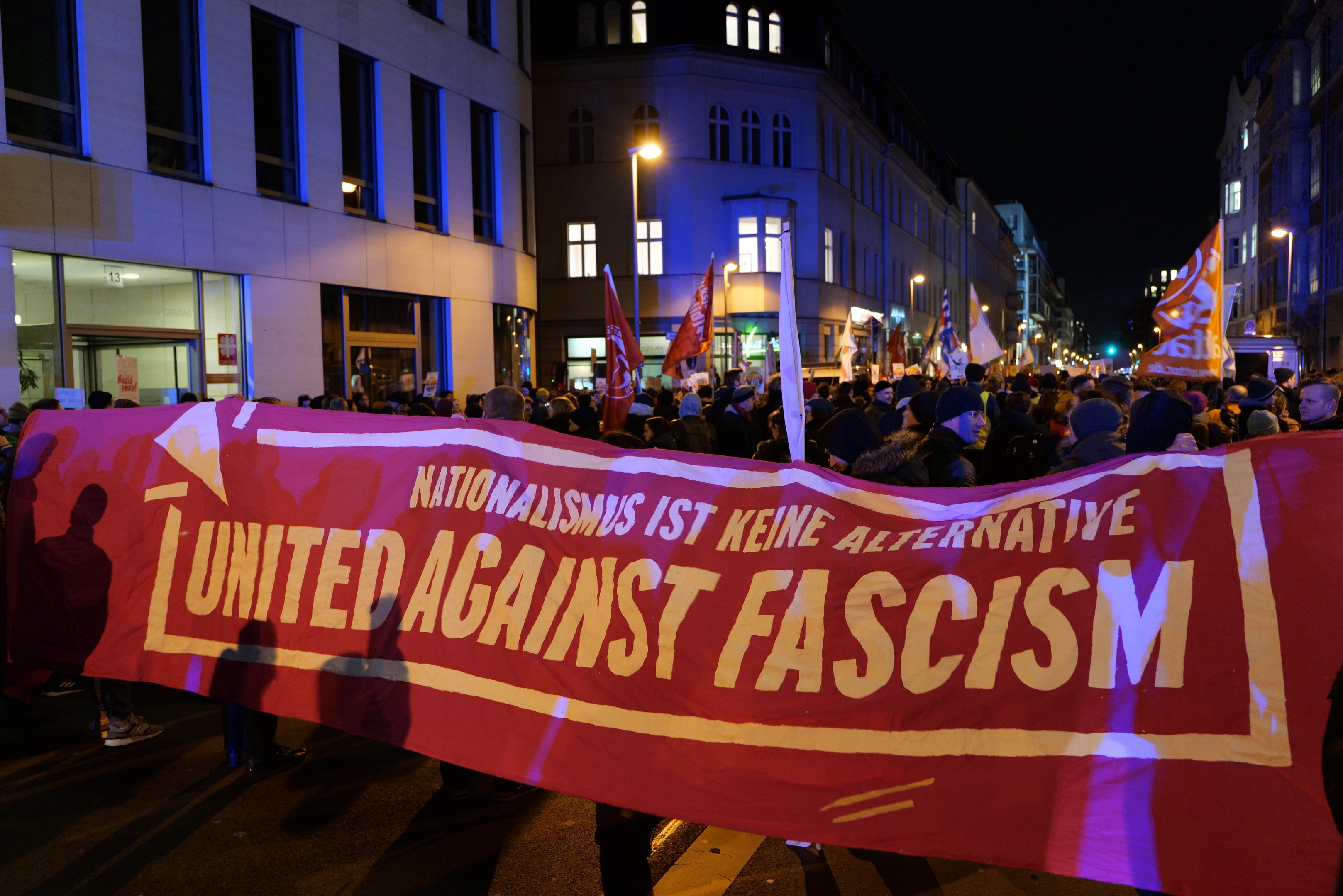 Eine Gruppe von Menschen marschiert auf einer Straße, die ein Banner mit der Aufschrift 'Einheit gegen Faschismus' trägt, mit Gebäuden, Laternenpfählen und Schildern an der Straße und dem Himmel im Hintergrund.