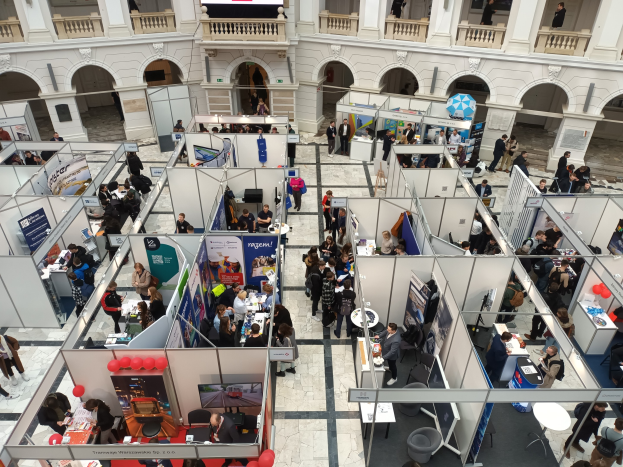 Eine große Gruppe von Menschen auf einer internationalen Fachmesse mit Ständen, Bannern, Tischen, Stühlen und Ballons in einem Raum mit architektonischen Bögen, Säulen und einem Bildschirm im Hintergrund.
