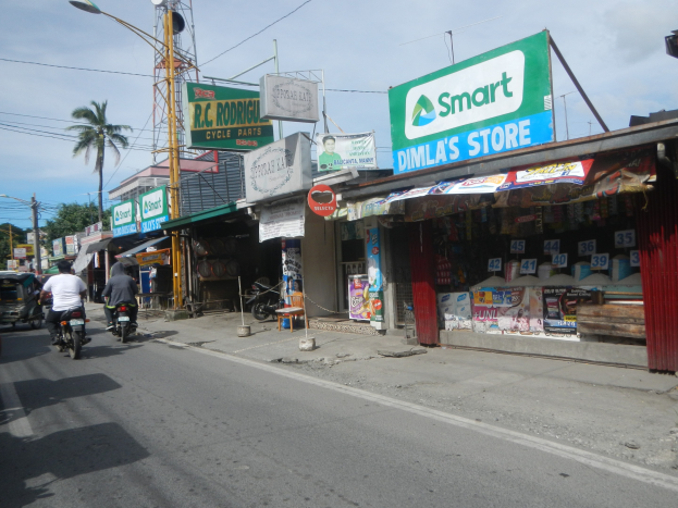 Eine belebte Straße mit Motorrädern, Autos, Fußgängern, Gebäuden, Schildern, Strommasten, Kabeln, Straßenlaternen, Bäumen und einem bewölkten Himmel.