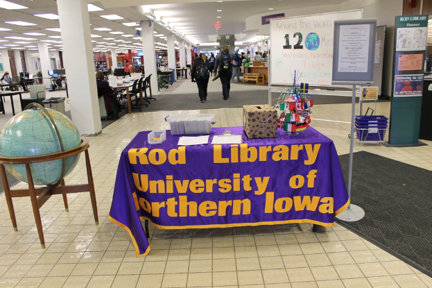 Eine Ausstellung mit einem Tisch, auf dem verschiedene Landesflaggen, eine Box, Papiere und eine Fahne sowie links daneben eine Weltkugel und rechts daneben eine Anschlagtafel mit einigen Menschen in der Nähe und mehrere Tische und Stühle im Hintergrund zu sehen sind.