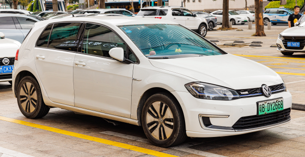 Ein weißer Volkswagen e-Golf auf einem Parkplatz neben anderen Autos geparkt, eine Person in der Nähe, Bäume, Gebäude und ein klarer blauer Himmel im Hintergrund.