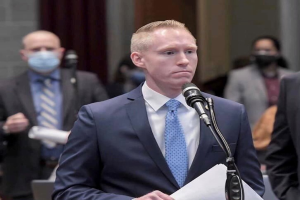 Ein Mann im Anzug und Krawatte spricht an einem Podium mit Papieren in der Hand, während einige Menschen hinter ihm in der Nähe einer Vorhangwand stehen.