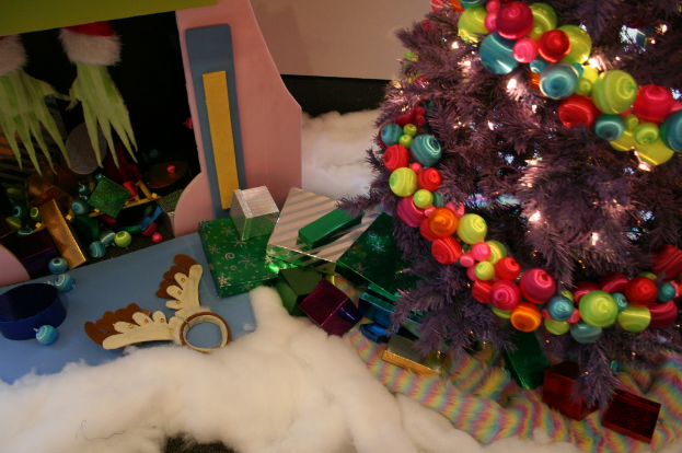 Drinnen steht rechts ein geschmückter Weihnachtsbaum mit Lichterkette und Schmuck, in der Mitte Geschenkboxen und Dekorationen, links oben eine Hütte mit Dekorationen und unten Wolle und Baumwolle.