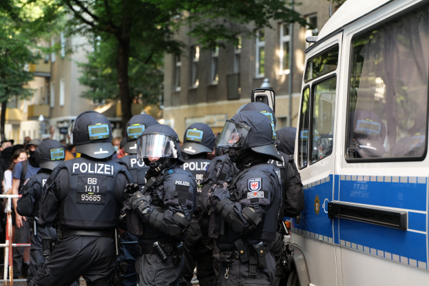 Eine Gruppe von Polizisten vor einem Bus auf einer Straße mit einer Menge, Absperrungen, Bäumen und Gebäuden im Hintergrund.
