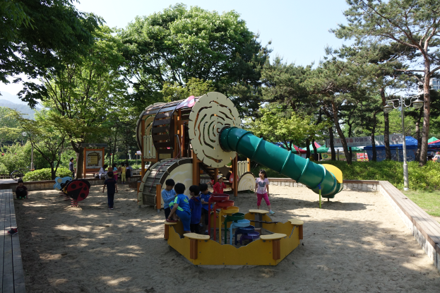 Eine Gruppe von Kindern spielt auf einem Spielplatz mit einer Rutsche in der Mitte, umgeben von Sand und Bäumen, mit einer hölzernen Plattform links daneben, Zelten, Laternenmasten und einem klaren blauen Himmel im Hintergrund.