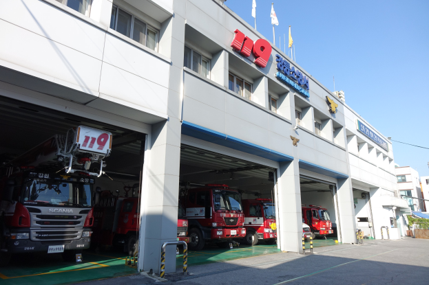 Feuerwache mit mehreren Feuerwehrautos vor dem Gebäude geparkt, mit Fenstern, Säulen und Namensschildern, mit Flaggen im Vordergrund und anderen Gebäuden gegen einen klaren blauen Himmel.