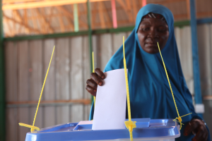 Frau in blauem Kopftuch mit einem Papier in der Hand an einer Wahlurne stehend, die ihren Stimmzettel in eine Box mit Stiften darin einwirft.