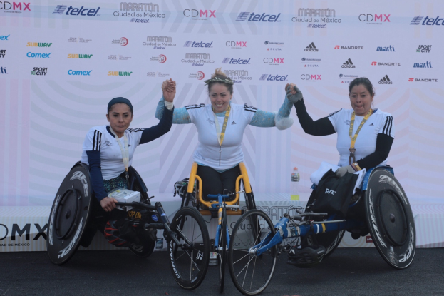 Drei Frauen im Rollstuhl auf einem Podium, die Hände erhoben, in weißen und schwarzen Kleidern und Medaillen, mit einem weißen Banner im Hintergrund.