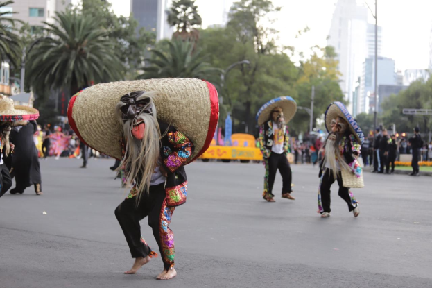 Eine Gruppe von Menschen in bunten Kostümen und Sombreros tanzt eine Straße in Mexiko-Stadt entlang, mit Bäumen, Gebäuden und Laternenpfählen im Hintergrund und einem sichtbaren Himmel.