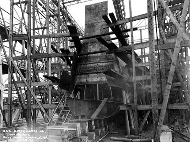 Schwarzes und weißes Foto eines Schiffs in einem Schiffbau, mit Holzpfählen und Treppen im Vordergrund und Himmel im Hintergrund, beschriftet mit "Marinebewaffnung" in der linken unteren Ecke.
