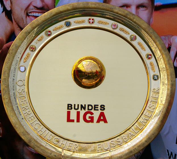 Mann hält goldenen Schild mit 'Bundesliga'-Text in der Hand, im Hintergrund eine Fahne mit drei Personen