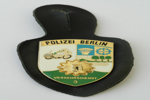Ein schildförmiges Polizewemzeichen mit der Aufschrift "Polizei Berlin" auf einem blauen Hintergrund, einem weißen Rand und einem zentralen weißen Stern, das auf einer Oberfläche liegt.