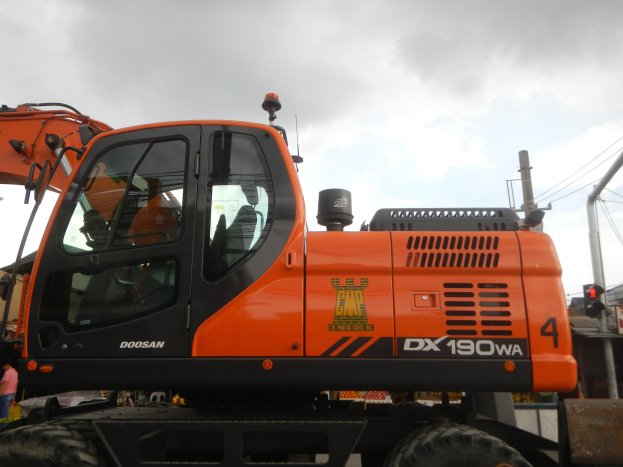 Oranger und schwarzer Doosan DX190WA Radlader vor einem Gebäude geparkt mit ein paar Menschen links daneben, Masten und Drähte im Hintergrund unter einem klaren blauen Himmel.