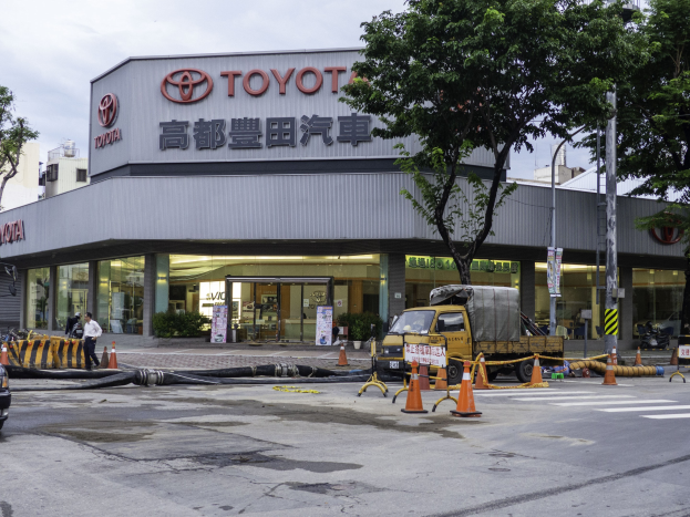 Ein Toyota-Autohaus in Tokio, Japan, mit Fahrzeugen, Verkehrskegeln, Rohren, Menschen, Bäumen, Pfählen, Planken und Gebäuden im Vordergrund und einem bewölkten Himmel im Hintergrund.