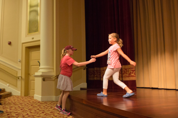 Zwei junge Mädchen tanzen auf einer Bühne vor einem Vorhang, mit einer Person, die auf einem Stuhl links von ihnen sitzt, einer Säule und einer Wand im Hintergrund.