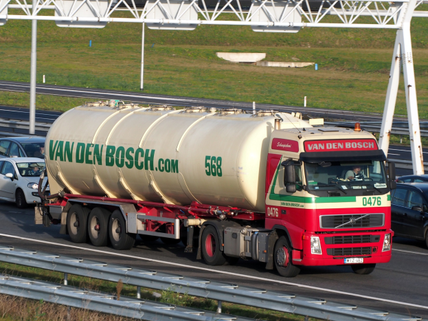 Ein großer weißer, roter und grüner Tanklastzug fährt auf einer Autobahn umgeben von anderen Fahrzeugen, mit einer Straßenbefestigung, Gras und einem Eisenrahmen im Hintergrund.