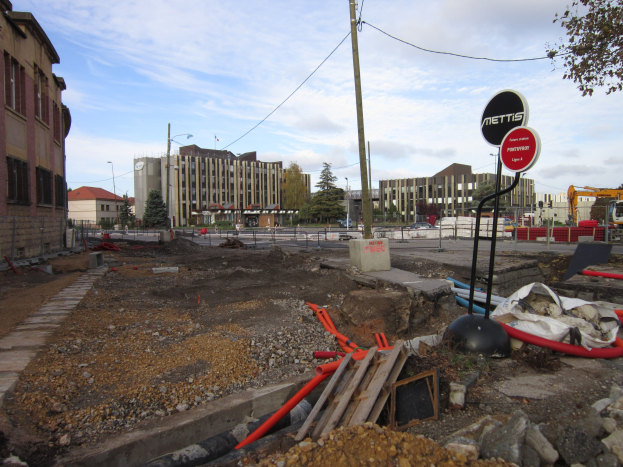 Baustelle mit verstreuten Materialien wie Steinen und Rohren, Schilder an Pfählen, ein Zaun, Fahrzeuge, Bäume, Gebäude mit Fenstern, Laternenpfähle und ein bewölkter Himmel im Hintergrund.