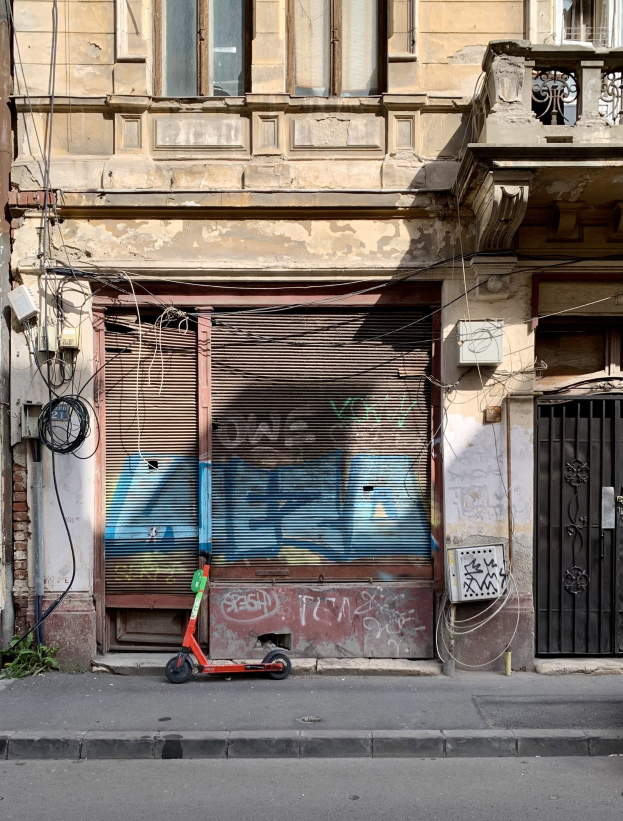 Ein roter E-Scooter auf einer städtischen Straße neben einem Gebäude mit Fenstern, Rollläden und einem Tor geparkt, mit sichtbaren Kabeln und Straßenobjekten und Text am unteren Rand.