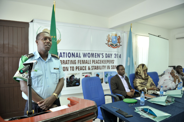 Ein Mann steht an einem Podium mit einem Mikrofon und Papier, während er eine Sitzreihe mit verschiedenen Gegenständen auf dem Tisch und Flaggen, einem Banner und anderen Elementen im Hintergrund anspricht.