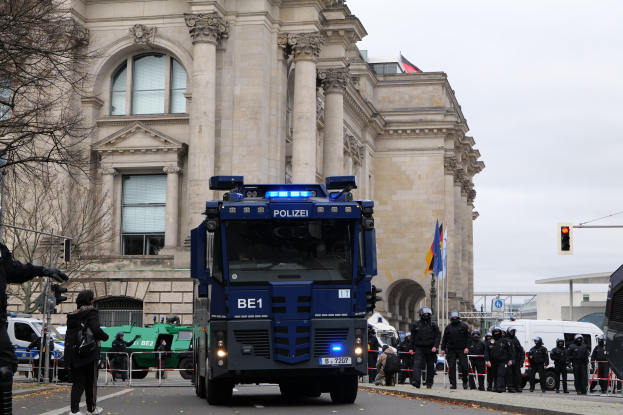 Gruppe von Polizeibeamten vor einem großen Gebäude mit architektonischen Details, Fahrzeugen auf der Straße, einer Person mit einer Kamera auf der linken Seite, Bäumen, Verkehrszeichen, Fahnenmasten und einem klaren blauen Himmel.