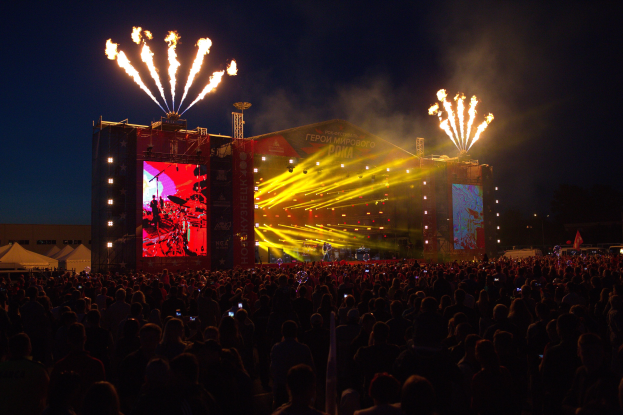 Eine große Menschenmenge auf einem Musikfestival mit einer Bühne, auf der Künstler unter hellen Scheinwerfern spielen, umgeben von Bannern und Zelten links und Feuerwerk am dunklen Himmel.
