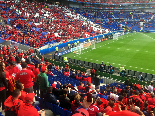 Ein volles Stadion mit Zuschauern, einige sitzen und einige stehen, die ein Fußballspiel mit Torpfosten, Bannern und Deckenbeleuchtung beobachten.